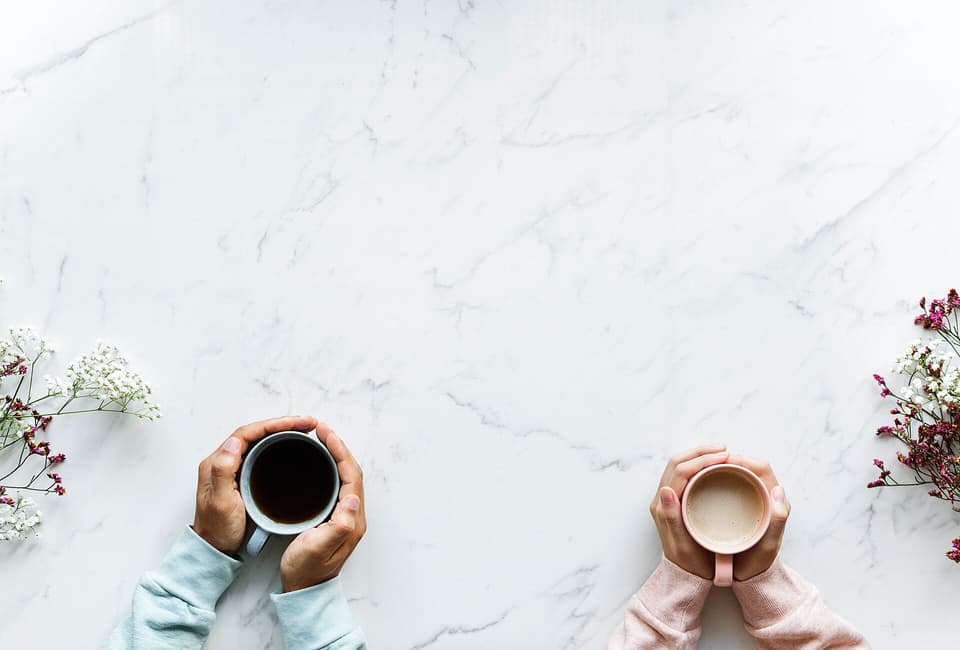 Two people’s hands holding warm drinks on a white marble table, seen from above (illustrative)