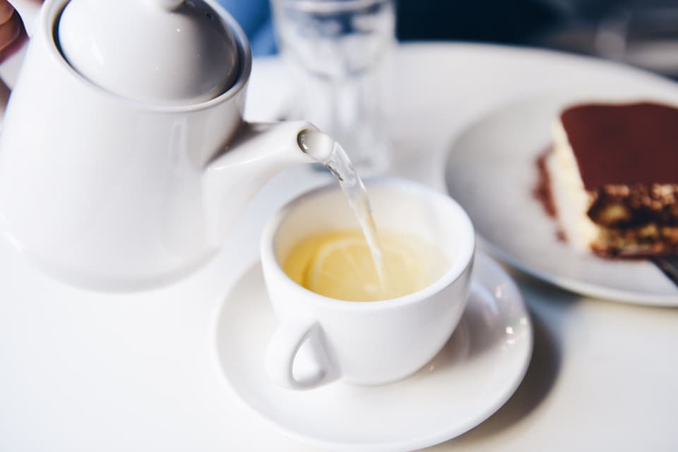 Tea being poured from a white teapot into a cup, beside a slice of cake (illustrative)