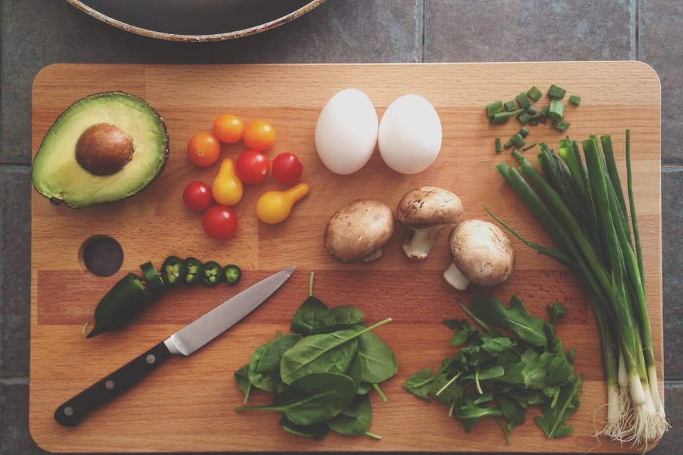 Avocado, eggs, mushrooms, tomatoes and greens on a wooden chopping board beside a pan (illustrative)