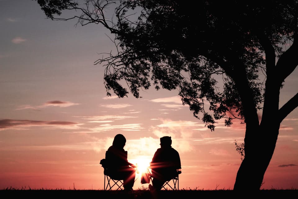 Two people sitting side by side in folding chairs under a tree, silhouetted against a pink sunset (صورة توضيحية)
