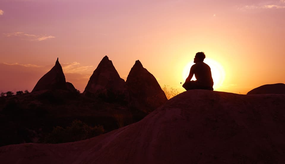 Distant silhouette of a person sitting cross-legged on a rock at sunset (illustrative)