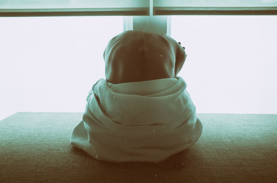 A person wrapped in a blanket sitting quietly before a bright window, seen from behind (illustrative)