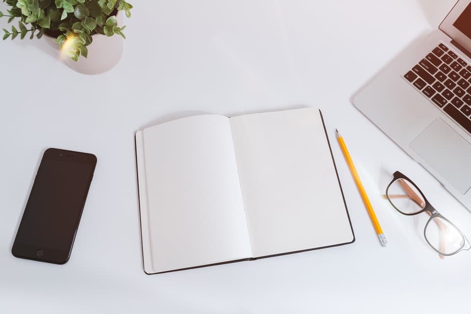 A tidy white desk with an open notebook, a pencil, glasses, a phone and a laptop, seen from above (illustrative)